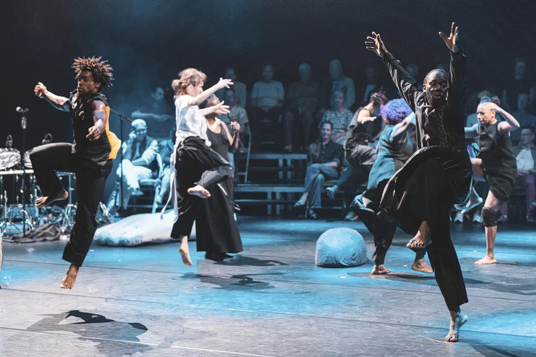 A group dressed in dark clothing dances and leaps across the stage. The stage is dark. The audience is seated in the background.
