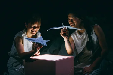 Die Performer:innen Takeshi Matsumoto und Makiko Aoyama sind in ein weiches Licht getaucht, halten gefaltete Papierfiguren über einem beleuchteten Kasten und lächeln.