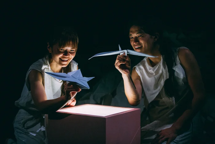The performers Takeshi Matsumoto and Makiko Aoyama are bathed in soft light and hold folded paper figures over an illuminated box, smiling.