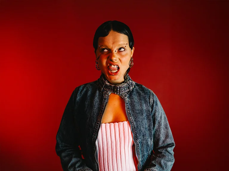 Rapper Marnele in front of a red background. Her facial expression is expressive and rebellious – she grimaces, showing her teeth, and looks slightly upward to the right.