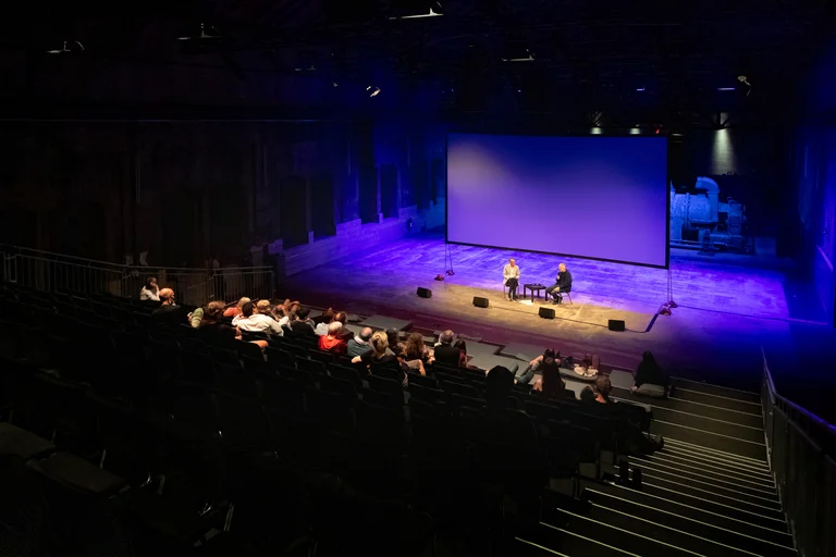 Audience in an industrial hall watching a conversation between Ivo Van Hove and Will Gregory on stage in front of a large screen.