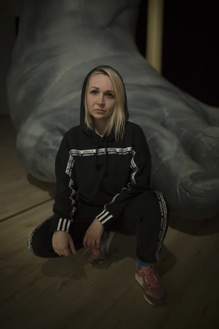 Anka Herbut, wearing a black Adidas tracksuit with white stripes, is kneeling on a wooden floor. She is wearing pink and white sneakers. In the background, an oversized art piece in the shape of a foot is visible. The scene is set in dim lighting.