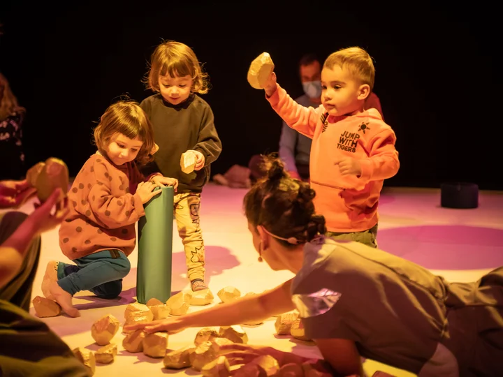 Szene aus der partizipativen Performance UNIVERS: Performer:innen und Kinder sitzen auf dem Boden und spielen gemeinsam mit kleinen, steinähnlichen Objekten.