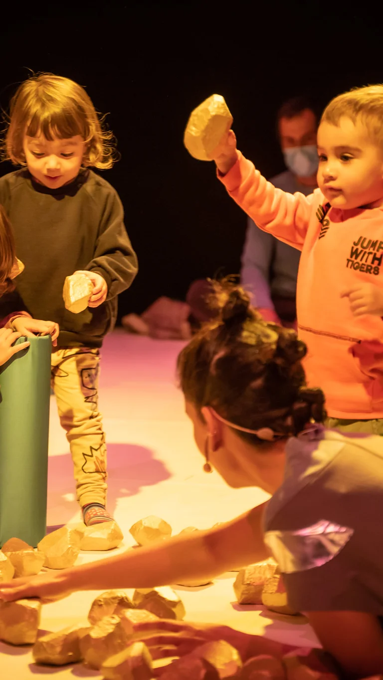 Szene aus der partizipativen Performance UNIVERS: Performer:innen und Kinder sitzen auf dem Boden und spielen gemeinsam mit kleinen, steinähnlichen Objekten.