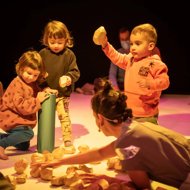 Szene aus der partizipativen Performance UNIVERS: Performer:innen und Kinder sitzen auf dem Boden und spielen gemeinsam mit kleinen, steinähnlichen Objekten.