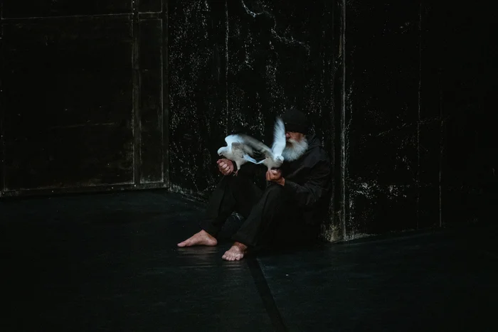 A man with a white beard sits barefoot in a dark stage corner, holding two white doves in his hands. The scene appears quiet, poetic, and full of symbolism.