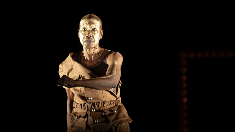 Germaine Acogny mit goldener Gesichtsbemalung in braunem Kostüm, ausdrucksstark vor schwarzem Hintergrund.