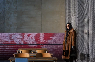 Eine Person steht seitlich an einer Bühnenwand neben mehreren Schulbänken. Sie trägt einen langen Mantel, Handschuhe und eine Sonnenbrille; der Raum ist klar ausgeleuchtet.