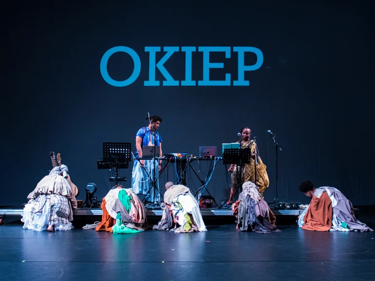 Fünf Performer:innen in bunten Kostümen knien und hocken vor zwei Musiker:innen auf einer Bühne. Im Hintergrund ist in großen blauen Buchstaben das Wort "OKIEP" zu lesen.