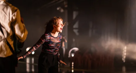 A person stands on a dimly lit stage with their mouth open and arms extended downward. Their hair is blown back. Another person is partially visible in the foreground, while spotlights shine through the hazy background.