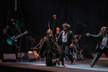 Three performers dance with expressive gestures on stage while a guitarist and a drummer play in the background.