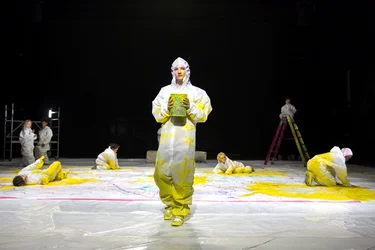 Eine Person in einem weißen Schutzanzug mit bunten Farbflecken steht in der Mitte der Bühne. Um sie herum sitzen und liegen Kinder, ebenfalls in Schutzanzügen, auf denen sich bunte Farbflecken befinden.