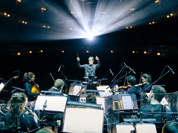 Conductor stands in front of an ensemble of musicians on a theatre stage; stage lights beam above, and music stands with scores are visible in the foreground.