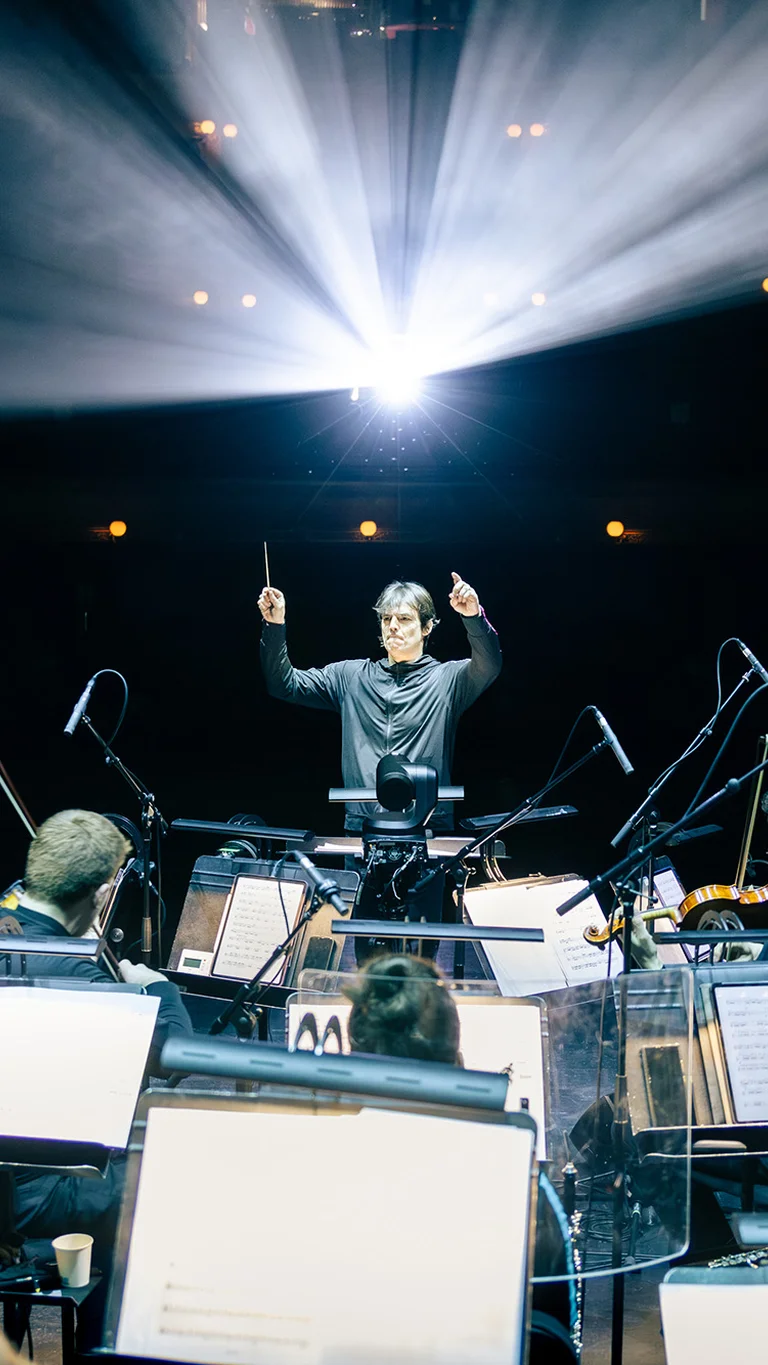 Dirigent vor einem Ensemble aus Musiker:innen auf einer Theaterbühne; über ihm strahlen Scheinwerferlicht und im Vordergrund sind Notenpulte mit Partituren zu sehen.