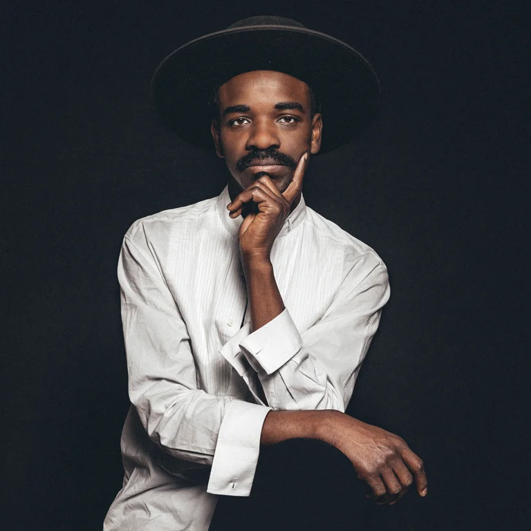Patrick Kabré is sitting in front of a black background and looking into the camera. He is wearing a white shirt and a black hat.