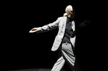 Anne Teresa De Keersmaeker in grauem Anzug bewegt sich seitlich über eine dunkle Bühne; der Hintergrund ist schwarz, die Choreografin wird von einem Spotlicht erfasst.