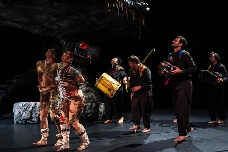 Two performers in colorful, imaginative costumes stand on stage looking upwards. Behind them, musicians play instruments including a drum, wind instrument, and accordion.