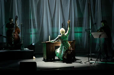 Joyce DiDonato sits at a wooden table on a dimly lit stage, wearing a white dress and raising one arm. To her left and right, musicians from Time for Three play double bass and violin. Long white fabric drapes hang in the background.