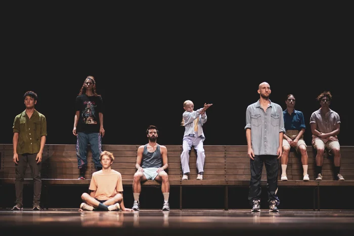 Performer:innen von "Futur Proche", die vor oder auf einer langen Bank sitzen oder stehen