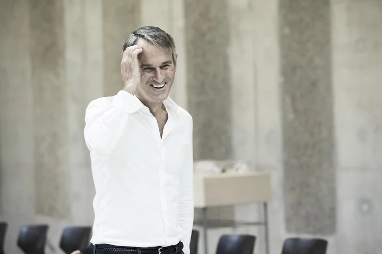 Ivo van Hove, artistic director of the Ruhrtriennale 2024-2026, wears a white shirt. He touches his forehead with his right hand and smiles.