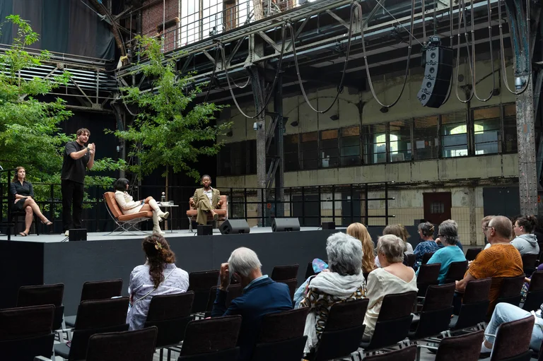 Fatima Khan and Jeremy O. Harris in the Jahrhunderhalle in Bochum. In the foreground of the picture you can see audience members looking at the stage. A sign language interpreter translates the conversation between the two protagonists