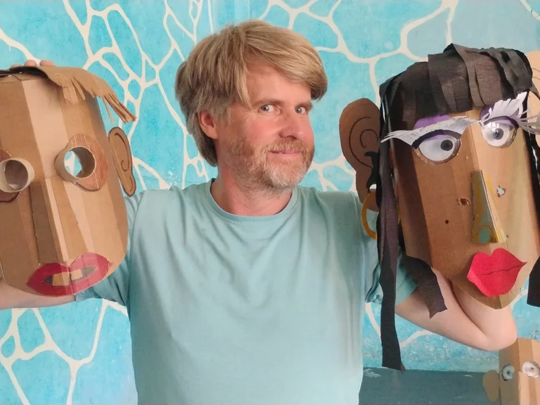 A person holds two large cardboard masks and looks into the camera.