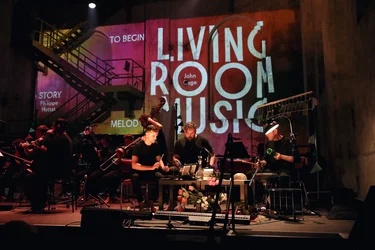 Musicians from the ensemble Les Apaches ! perform on stage in an industrial hall. In the foreground, three percussionists sit among various instruments and everyday objects, with string and wind players behind them. The words “LIVING ROOM MUSIC” and “John Cage” are projected in large letters onto the wall in the background.