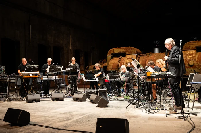 The Will Gregory Moog Ensemble is performing on a stage inside an industrial hall, with an old turbine in the background. Eleven musicians play electronic instruments, including synthesizers and mixing consoles. The stage is bathed in warm light, creating an atmosphere that blends technical precision with artistic expression.