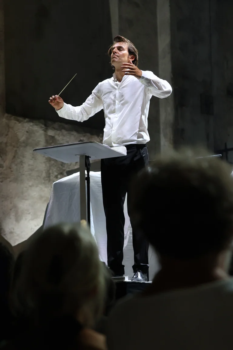 Conductor Julien Masmondet stands at the conductor’s podium with his arms raised, holding a baton in his right hand. He is wearing a white shirt and black trousers. The heads of audience members are visible in the foreground.