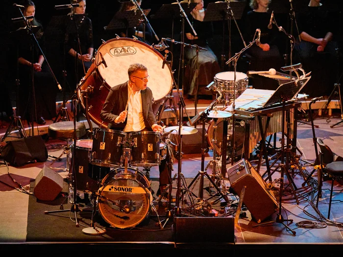 David Cossin of the Bang on a Can All-Stars on drums on stage, with members of Chorwerk Ruhr in the background.