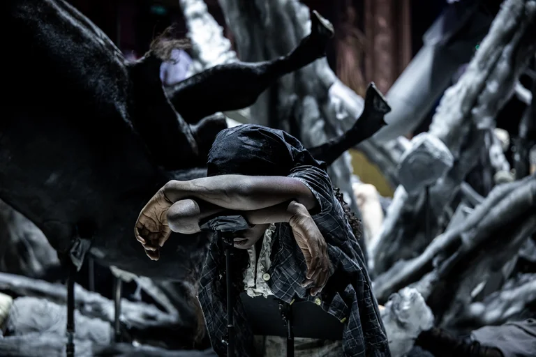 Eine Schauspielerin beugt sich erschöpft mit verschränkten Armen und gesenktem Kopf nach vorne. Hinter ihr ragen chaotisch geformte Kulissenteile und Skulpturen in Form von Körperteilen hervor. Die Atmosphäre ist bedrückend und düster.
