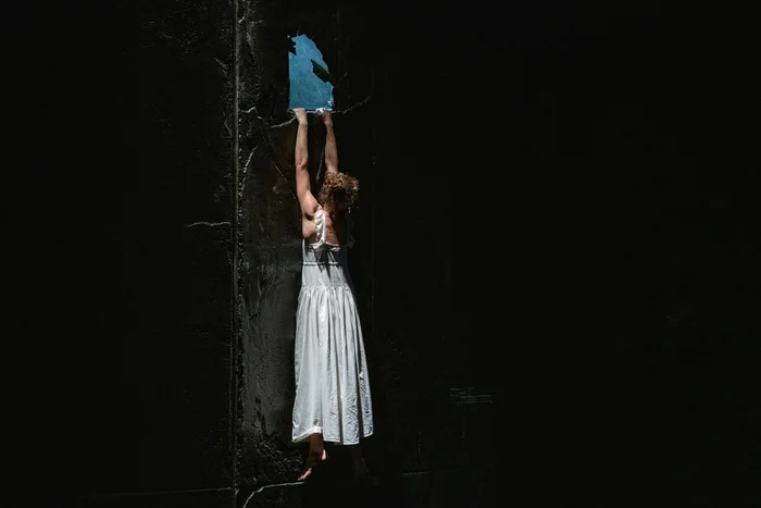 A woman in a white dress hangs from a broken hole in a dark wall, through which blue sky is visible.