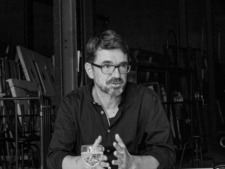 A black-and-white photograph of the dramaturg Bernd Isele sitting at a table, his arms resting on the table and his hands raised.