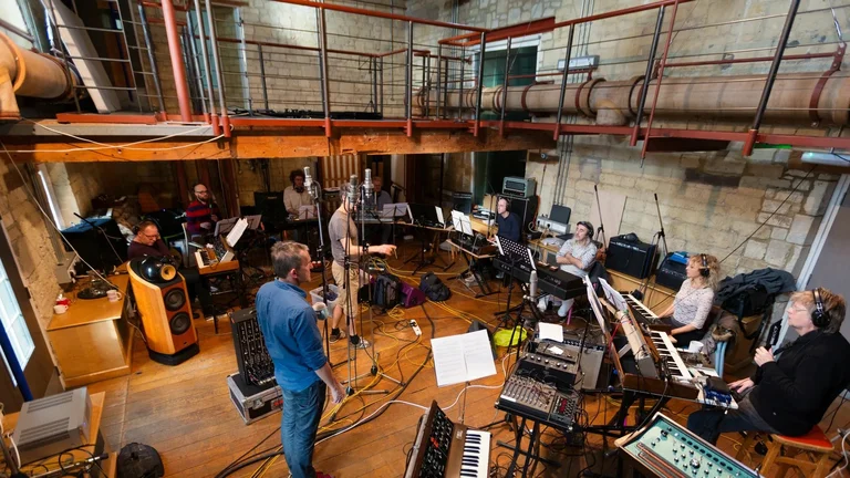 Das Will Gregory Moog Ensemble bei einer Probe in einem Raum mit rustikaler Steinwand und Holzboden. Die Musiker:innen sind in einem Halbkreis um eine Sammlung von Synthesizern, Notenständern und Mikrofonen angeordnet. Eine erhöhte Galerie ist im Hintergrund sichtbar.