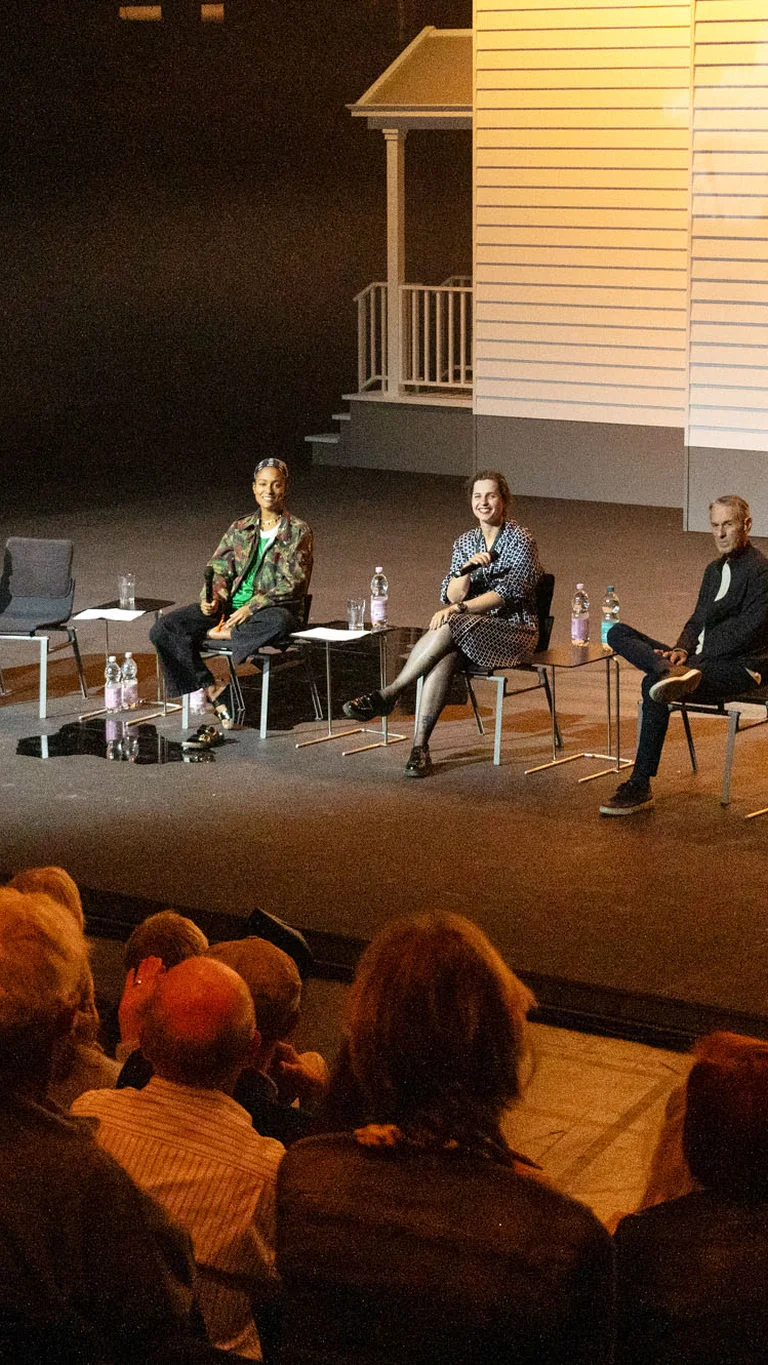 Public discussion with participants in the production ‘I Did It My Way’, who are seated in front of a white façade, the stage set. In the foreground, a blurred audience can be seen.