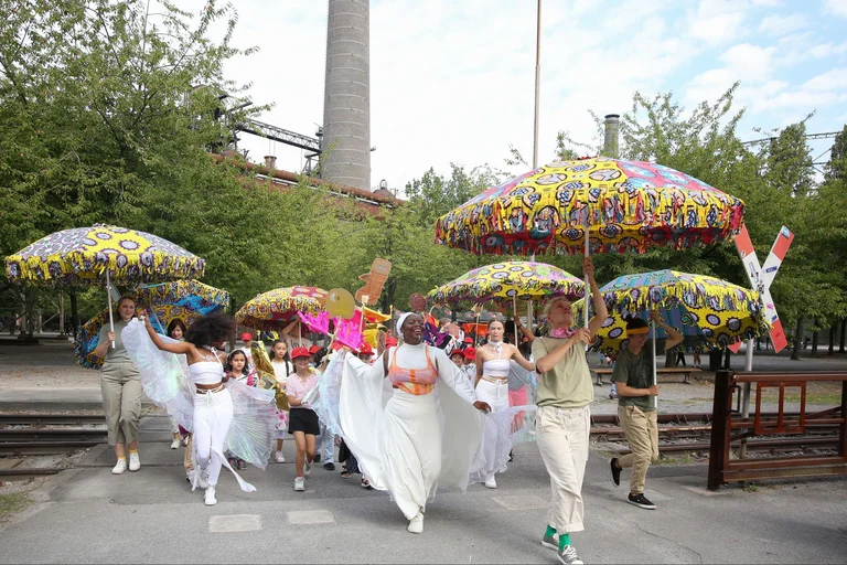 The ‘Celebration Parade’ marches through the Duisburg Nord Landscape Park. Colourful parasols are held in the air, people wear white clothing. The atmosphere is exuberant
