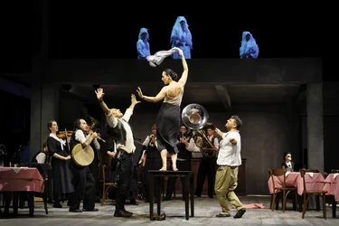 Scene from Balkan Erotic Epic: A dancer stands on a table, waving a cloth, surrounded by performers and musicians in a lively, ritual-like scene. Several white-clad figures appear on an upper level.