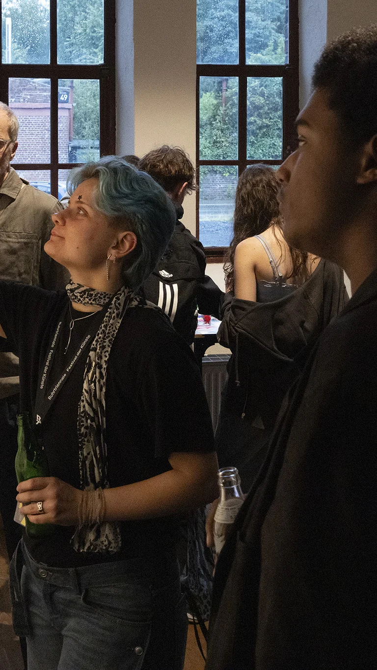 Eine Gruppe junger Menschen in einem Ausstellungsraum. Eine Person mit blauen Haaren deutet auf ein Bild an einer Wand.