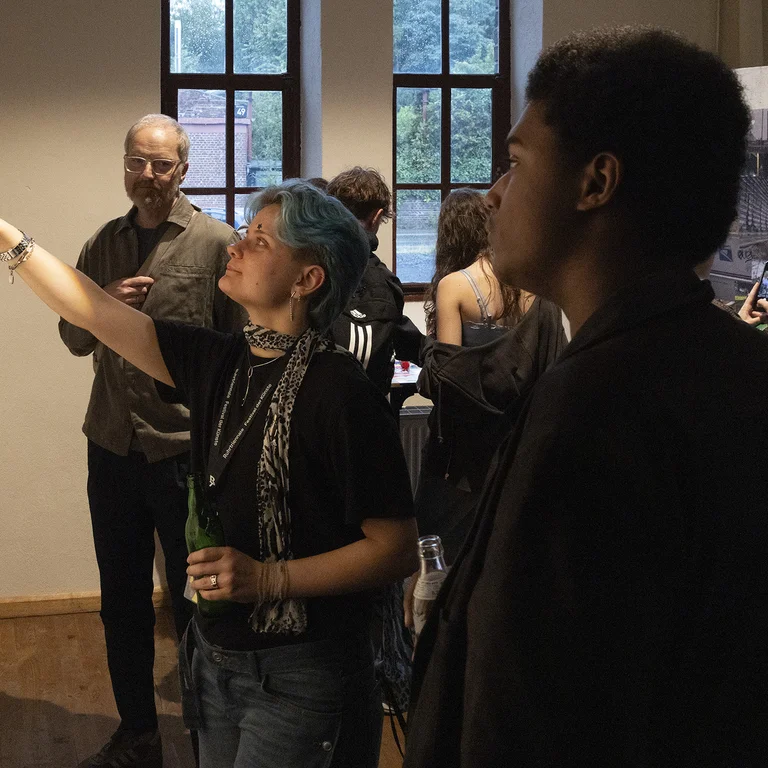 A group of young people in an exhibition space. A person with blue hair is pointing towards a picture hanging on a wall.