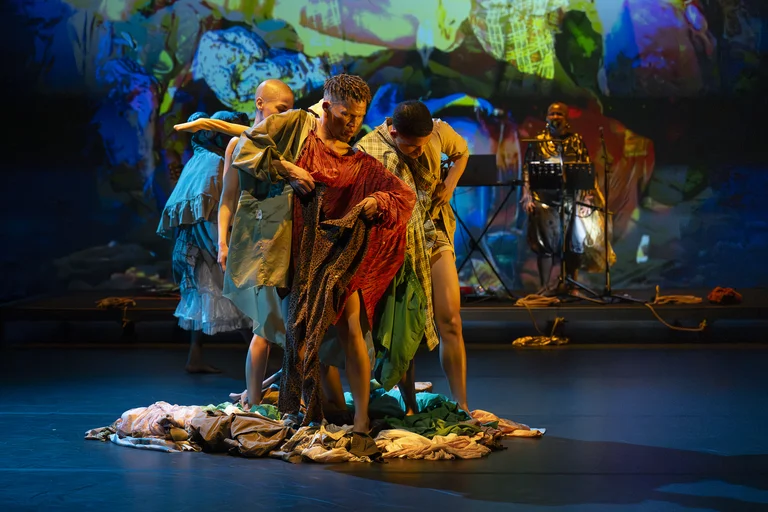 Performers stand closely together on a pile of clothes, wearing colorful costumes, with a vibrant projection in the background.