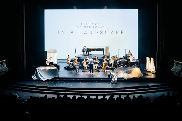 The ensemble Les Apaches ! performs on a large stage in front of an audience. In front of a bright projection displaying the words “In a Landscape,” a conductor leads the ensemble while a pianist sits at a grand piano on an elevated platform and other musicians perform across the stage.