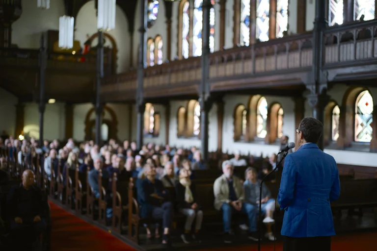 View inside the Evangelical Church at the Market in Katernberg during the event 124 Years of Reverb. In the foreground, a woman in a blue jacket speaks into a microphone facing the audience. The pews are filled with attendees, while colorful light streams through the large stained-glass windows.