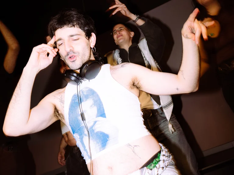 Hassandra mit Kopfhörern um den Hals steht hinter einem DJ-Pult und tanzt. Im Hintergrund feiern weitere Menschen.
