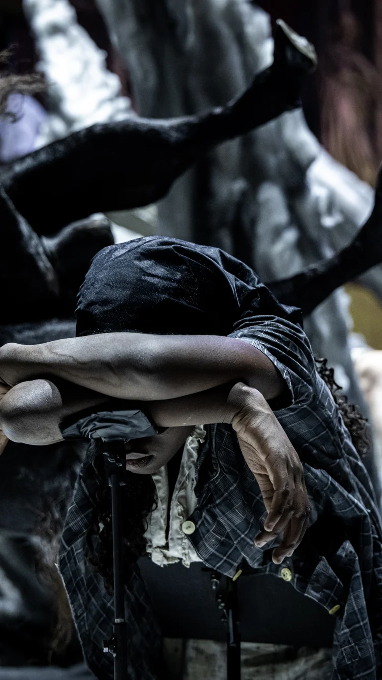 Eine Schauspielerin beugt sich erschöpft mit verschränkten Armen und gesenktem Kopf nach vorne. Hinter ihr ragen chaotisch geformte Kulissenteile und Skulpturen in Form von Körperteilen hervor. Die Atmosphäre ist bedrückend und düster.