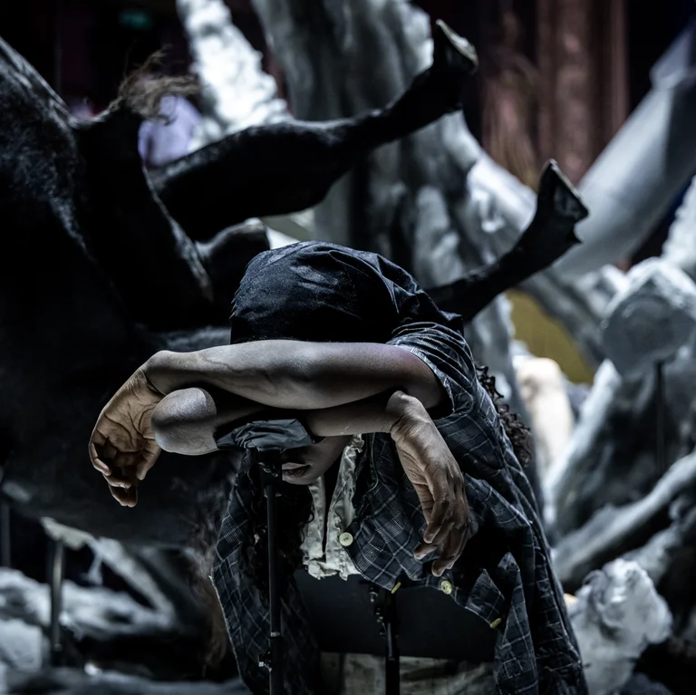 An actress bends forward in exhaustion with crossed arms and her head lowered. Behind her, chaotically shaped set pieces and sculptures in the form of body parts rise up. The atmosphere is oppressive and dark.