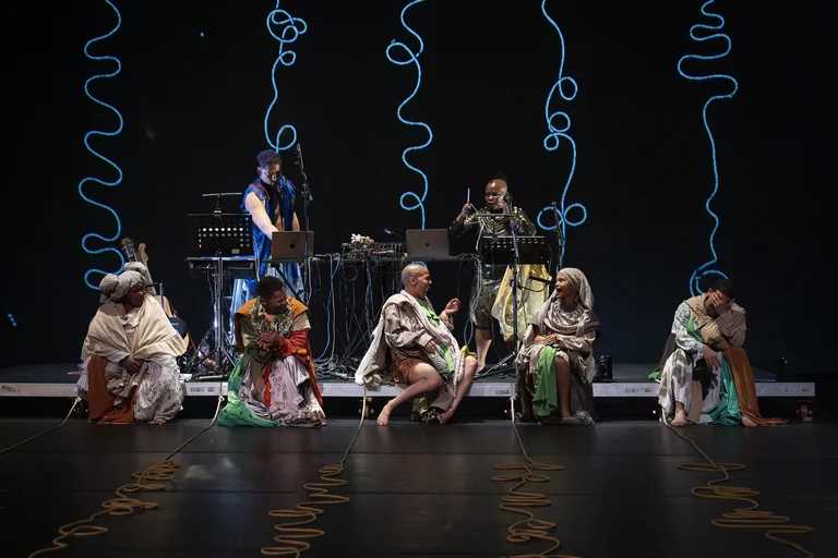 A stage performance with five seated performers in colorful robes and two musicians at electronic instruments in the background, with glowing blue lines projected behind them.