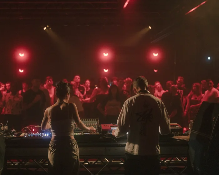 Two DJs stand behind their booth on a stage, with a crowd dancing and standing in front of them under red lights.