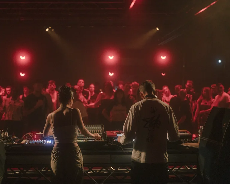 Two DJs stand behind their booth on a stage, with a crowd dancing and standing in front of them under red lights.