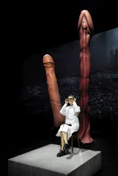 Scene from Balkan Erotic Epic: A person in white clothing sits on a platform, framed by two oversized penis sculptures. The lighting creates a surreal and symbolic atmosphere.