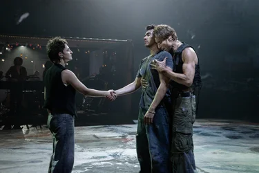 Three performers stand on a stage. Two people are positioned centrally, one directly behind the other; in front of them stands another person holding the hand of the performer opposite them.